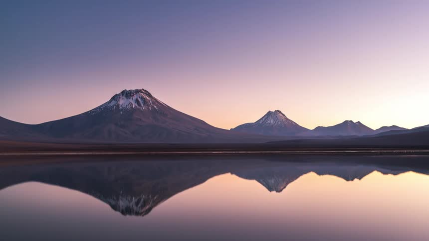 Landscape of a reflective lake surrounded by snowcapped mountains with the starry night transitioning into dawn in a timelapse