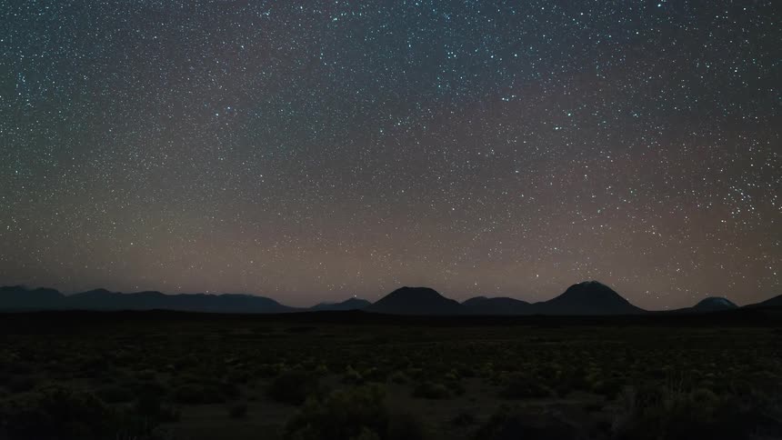 A desert region with mountains on the horizon and it is getting dark and the sky is full of stars and after a while it dawns