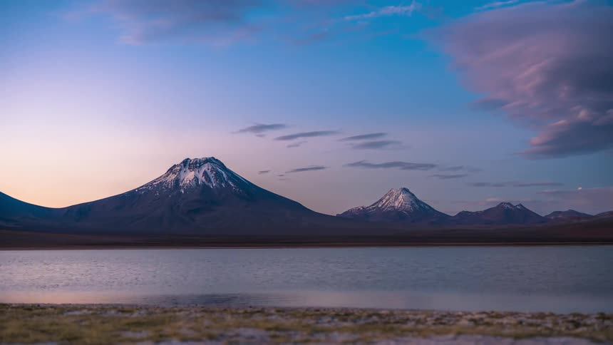 A region with several high mountains in front of a lake and it is getting dark