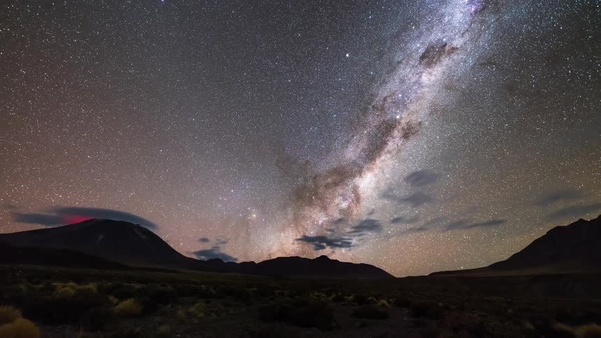 A desert region with mountains and in the sky there are many stars and a galaxy