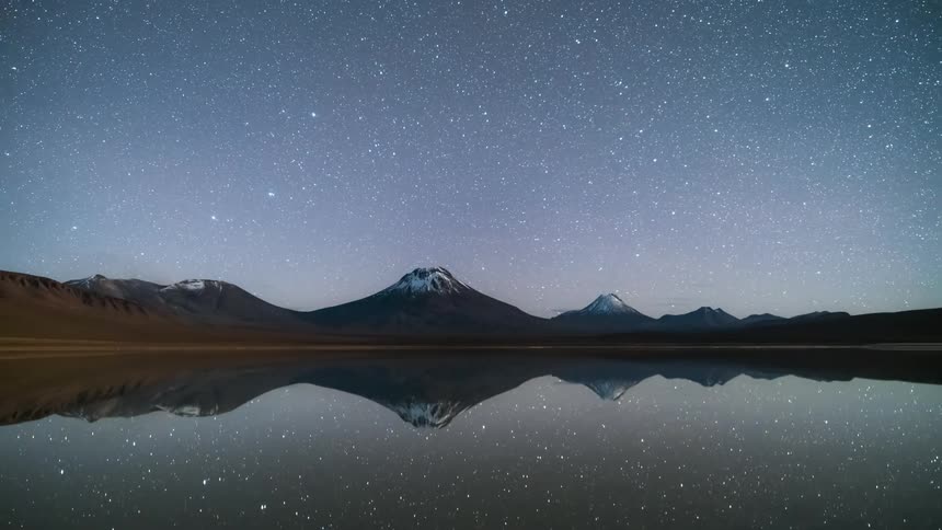 Mountains in front of the water that is reflecting them and the sky is full of stars and clouds appear