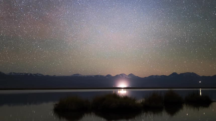A starry sky in a mountainous region has some vehicles passing close to the water that is reflecting the sky