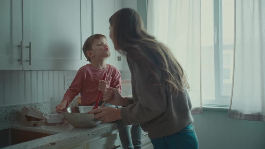A woman in a kitchen cooking with a young boy sitting on the kitchen counter playing at helping her then they kiss