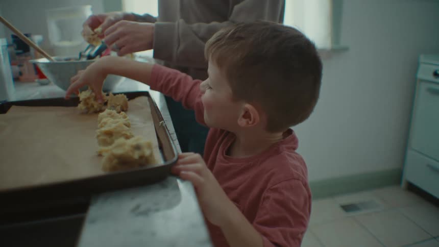 Boy helps his mother place pastries on cooking sheet then licks his fingers