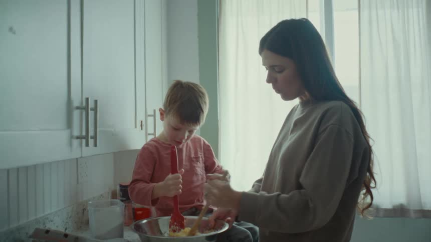 A woman standing with her son sitting at the counter and they are both preparing food and are happy