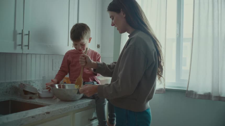 A mother and young son stir something in a bowl at the kitchen counter