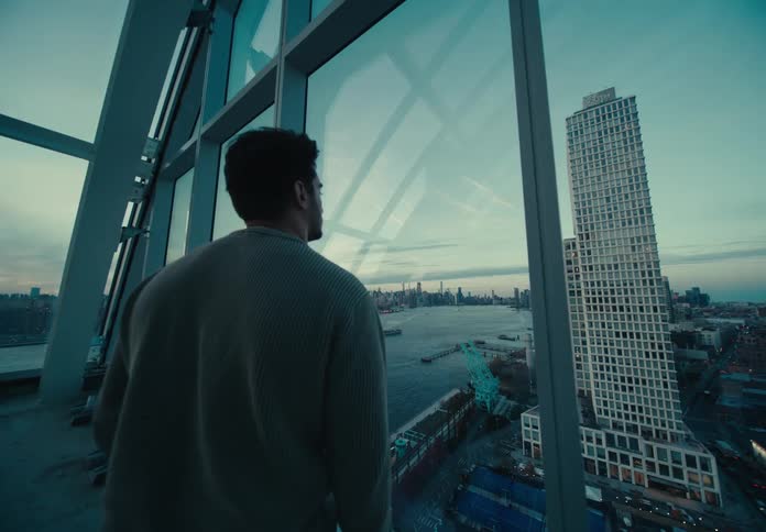 Man stands in glass room looking at river and the surrounding urban landscape