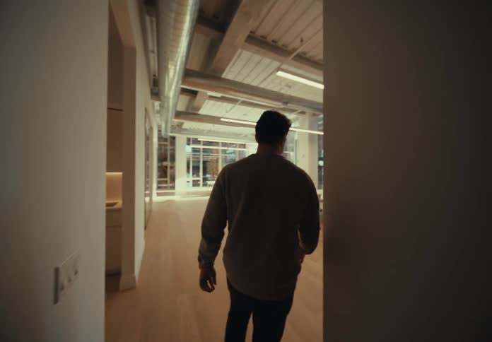 A person with his back turned is walking in a room that has just a table with some chairs a wooden floor and glass windows