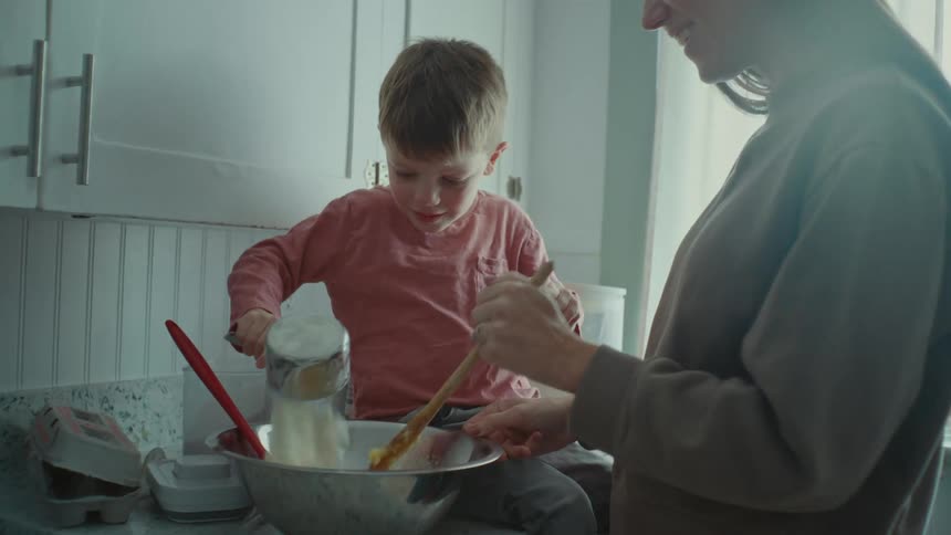 Mother and son add flour into a silver bowl and kiss each other in the kitchen
