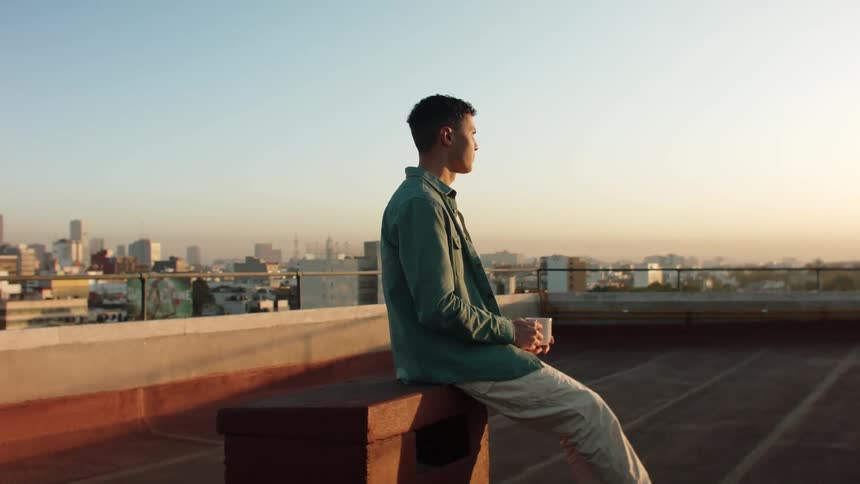 Man sitting on the terrace of a building overlooking a metropolis in the morning sky and drinking a cup of coffee
