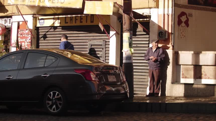 A man stands against the wall at midday while another man walks by on the sidewalk