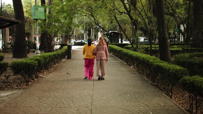 Two women are back to back and walking holding each others arms on a path surrounded by trees in a city square