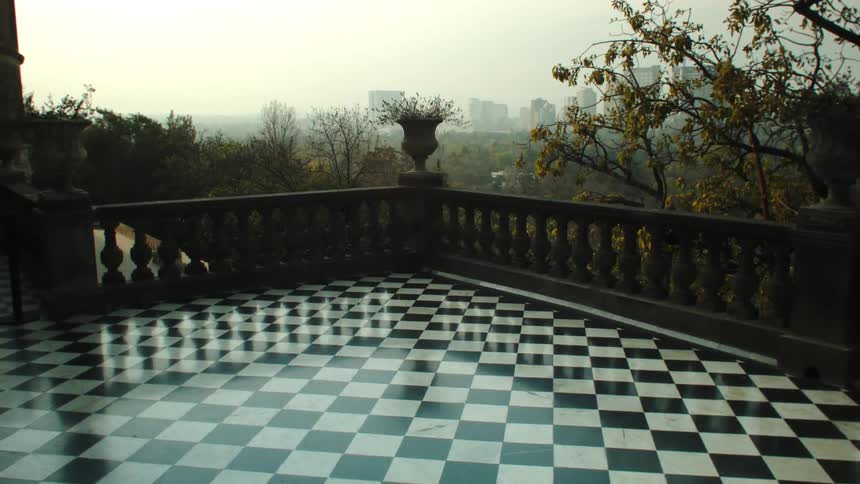 A black and white checkered floor and a small hollow concrete wall and on the horizon there are many trees and a city