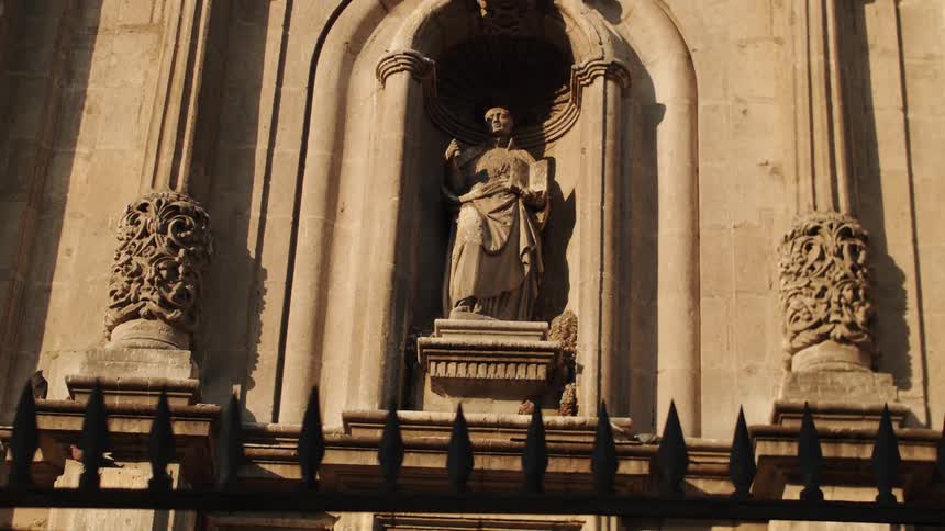 A statue in an alcove on a stone wall with carvings and a fence