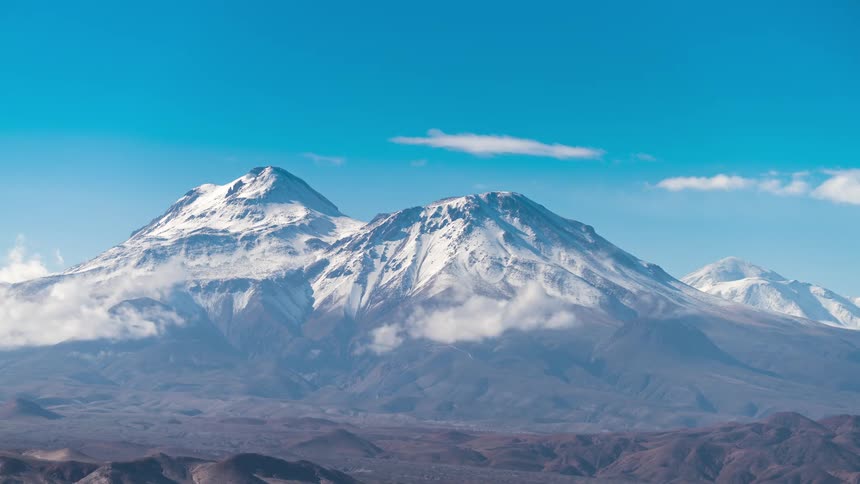 Clouds pass across snow covered mountains towering over hilly terrain