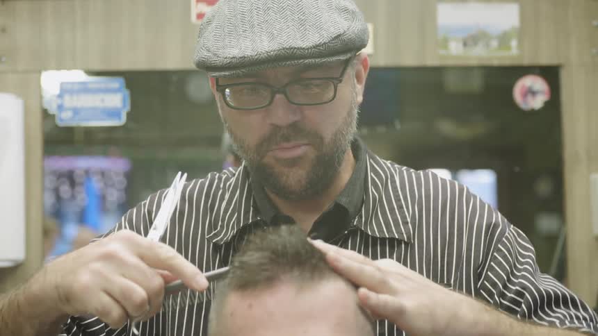 A barber in a hat smiles and talks while he works