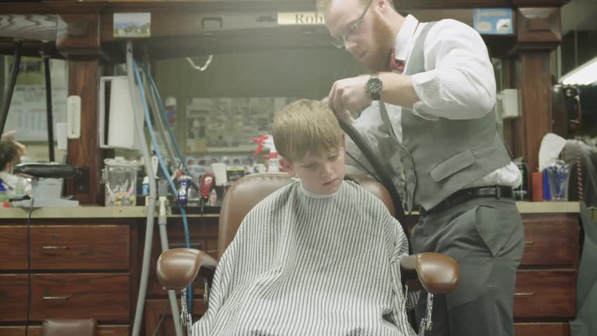 A boy sits in a barber chair while a barber cuts his hair