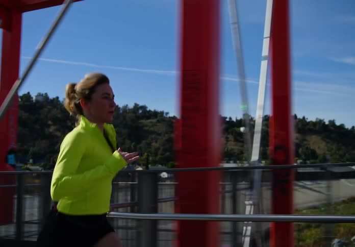 A woman in a bright yellow top is jogging on a bridge with metal railings