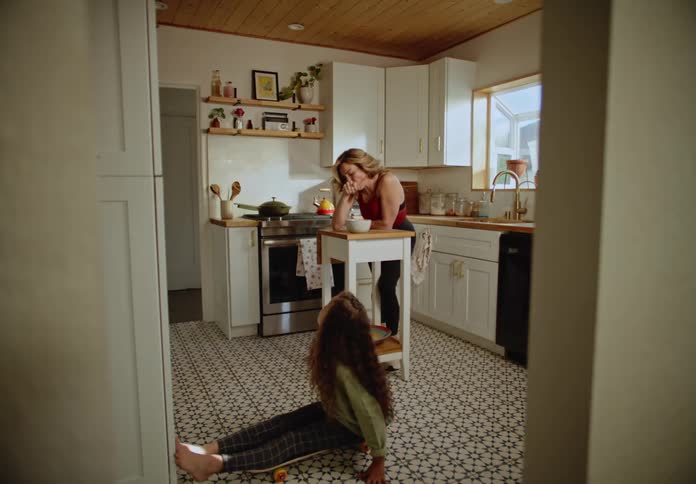 A mother eats cereal while her daughter plays on the kitchen floor