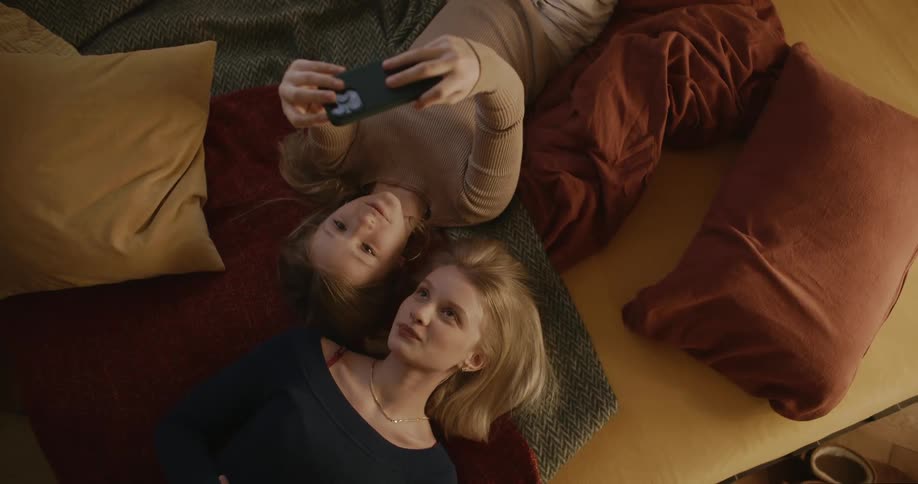 Two people lying on a rug with cushions taking selfies with a smartphone