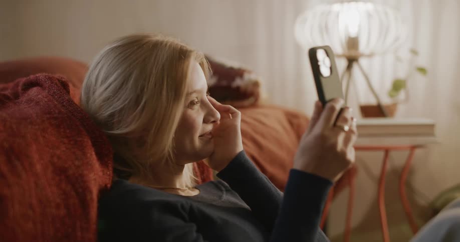 Young blonde woman sitting next to the bed laughing a lot while using her cell phone