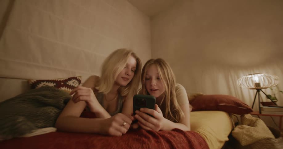 Two teenage girls over a bed with orange blankets looking at a phone