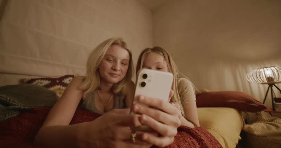 Two friends resting in bed looking at a smartphone that one of them is holding