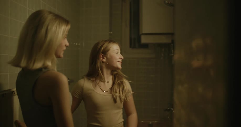 Young woman applying makeup to a teenager in the bathroom and then the girl checks how it looks on her cell phone