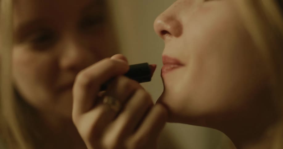 Female teen applies lipstick on another teens lips while smiling inside the restroom
