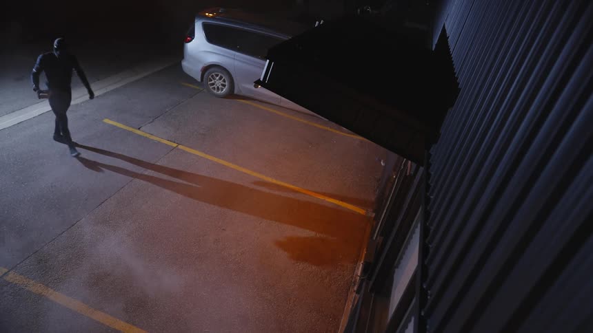 Masked man arriving at the facade of an establishment and throwing a stone at night