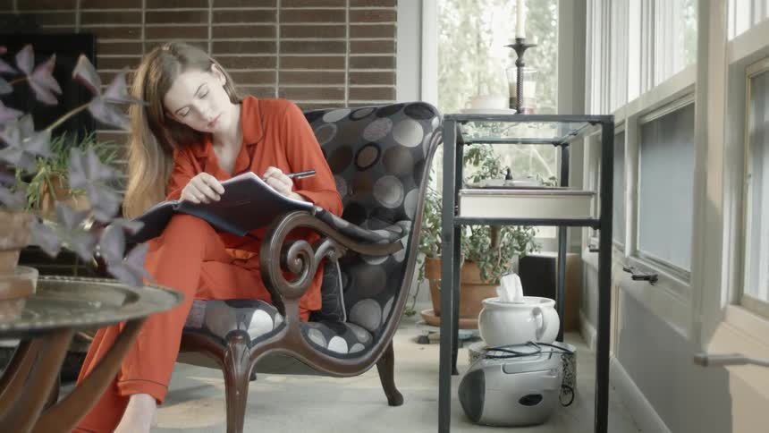 Woman sits in a chair next to a window and writes in a planner