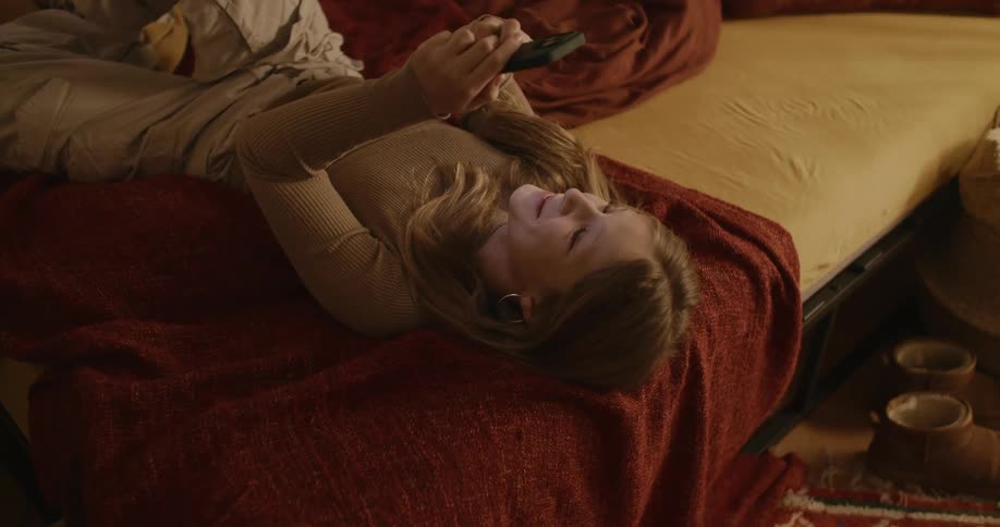 Blonde teenager rolling around in bed and using her cell phone smiling