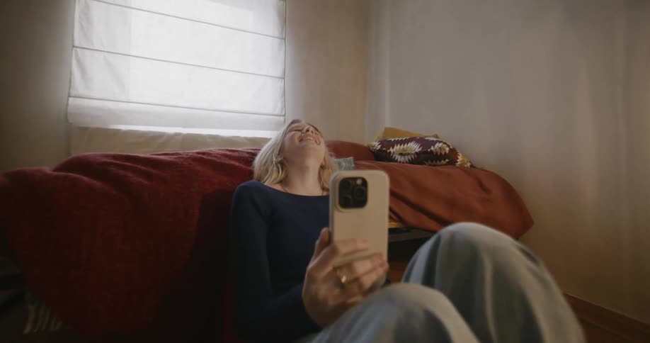 A blonde girl sitting on the floor of the bed is smiling while playing on her cell phone