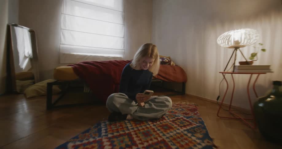 Young blonde woman sitting next to the bed on the floor and using her cell phone