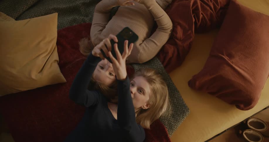 Two ladies taking selfies with a smart phone while laying on the floor indoor