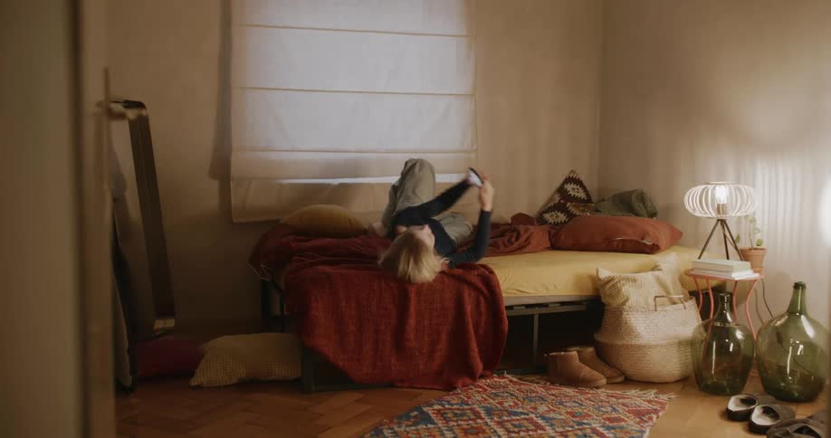 A woman rolls on her bed while looking at her cell phone