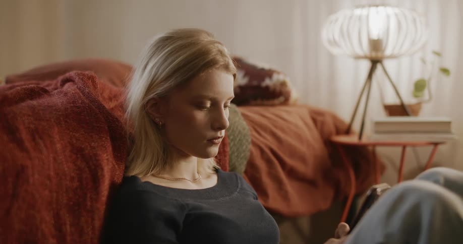 Portrait of a young blonde woman sitting next to the bed with a bored facial expression playing on her cell phone