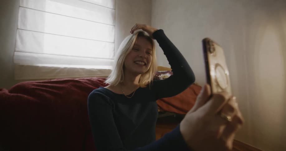 A person is taking a selfie in a room with a smartphone near a window and a bed