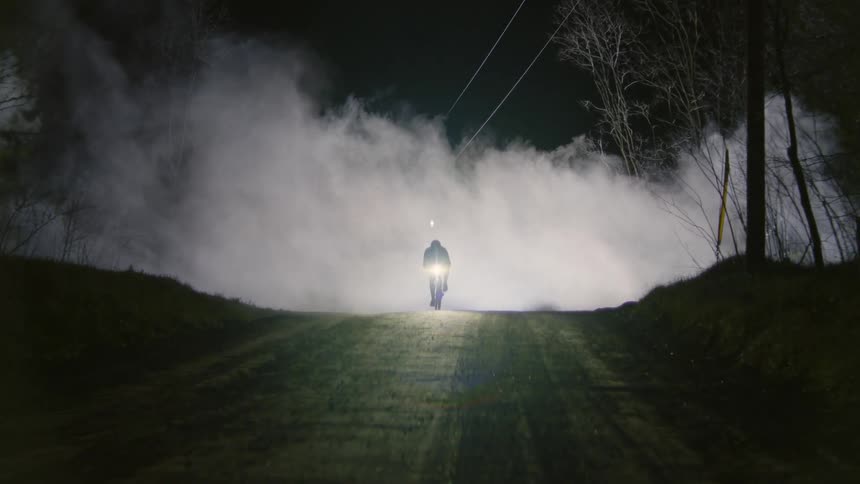 A large mysterious white smoke and a person riding a bicycle appears with a flashlight in front of the bike on a sandy street