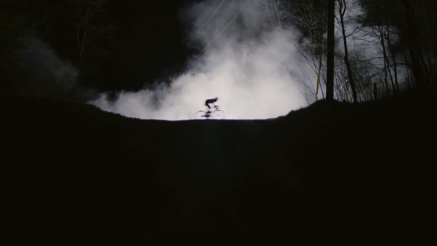A person on a bicycle speeding through a dark foggy forest at night