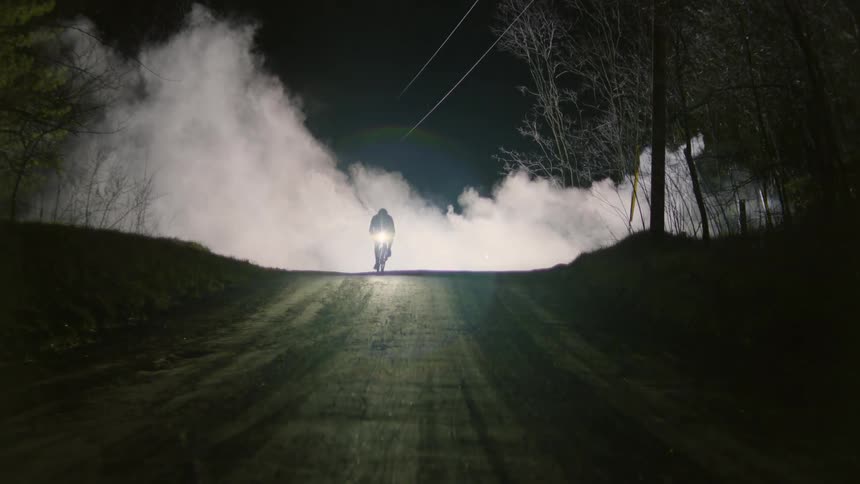 A cyclist is coming out of white smoke on a dirt street at night