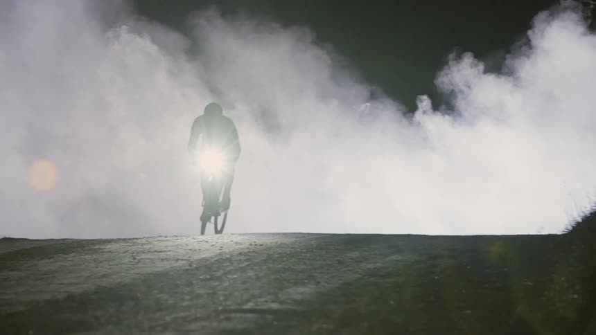 A man riding a bicycle comes out of the fog covering a road at night