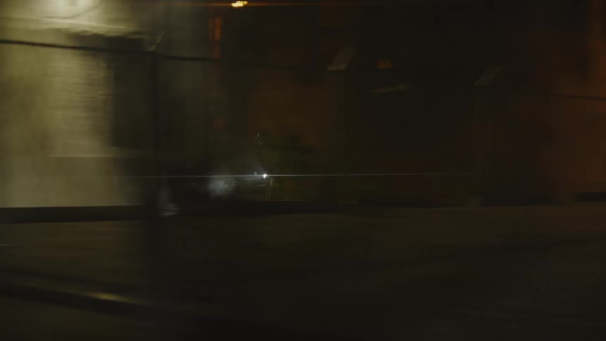 A person passing quickly on a bicycle near a parking lot with several bars on a parked car and it is at night