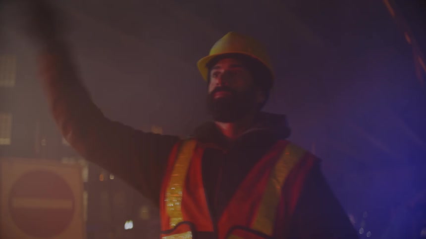 Worker directing traffic at night looks back at vehicle with flashing blue and red lights
