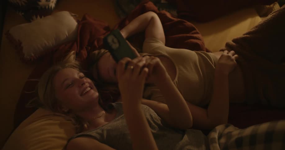 Two female teens lay on the bed smiling while looking at the smart phone being operated by one of them