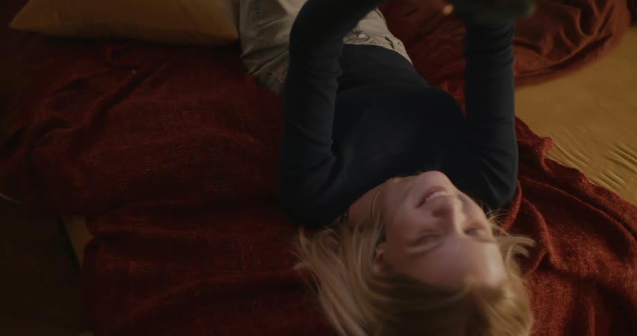 Woman rolls over onto her back on bed while smiling at smartphone