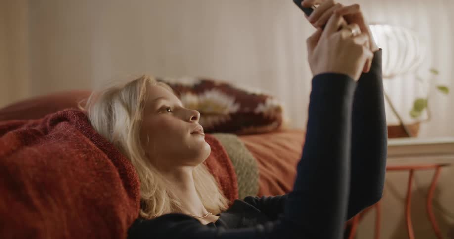 A teenage girl sitting on the floor by her bed adjusting her hair to take a selfie