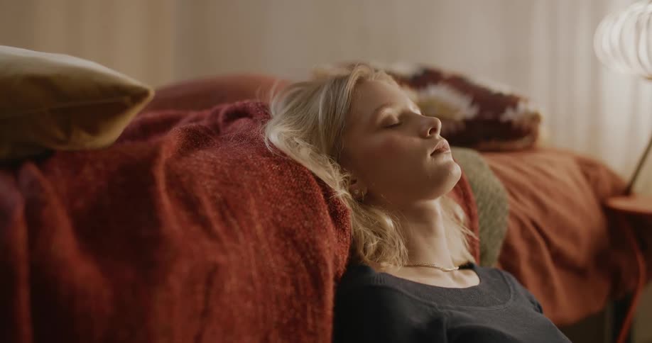 A blonde woman sits by a bed and rests her head on sheets