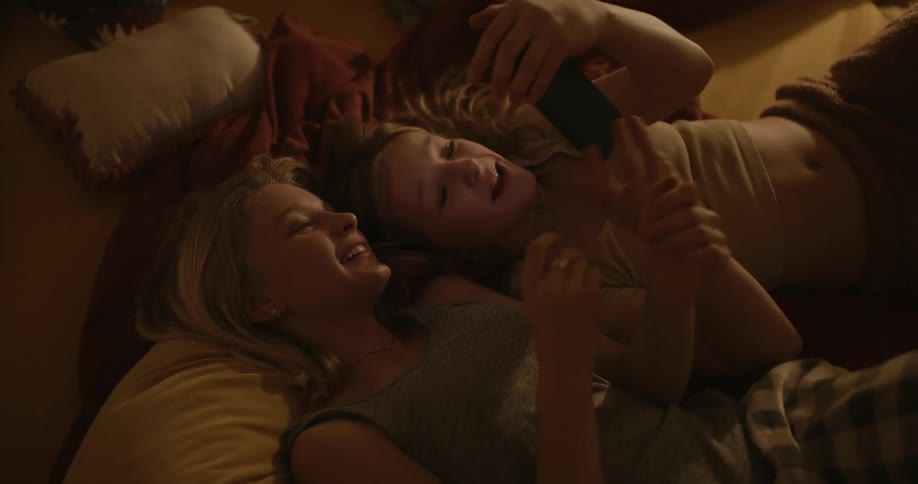 Two teenage girls lying on a bed talking and laughing at their smart phone
