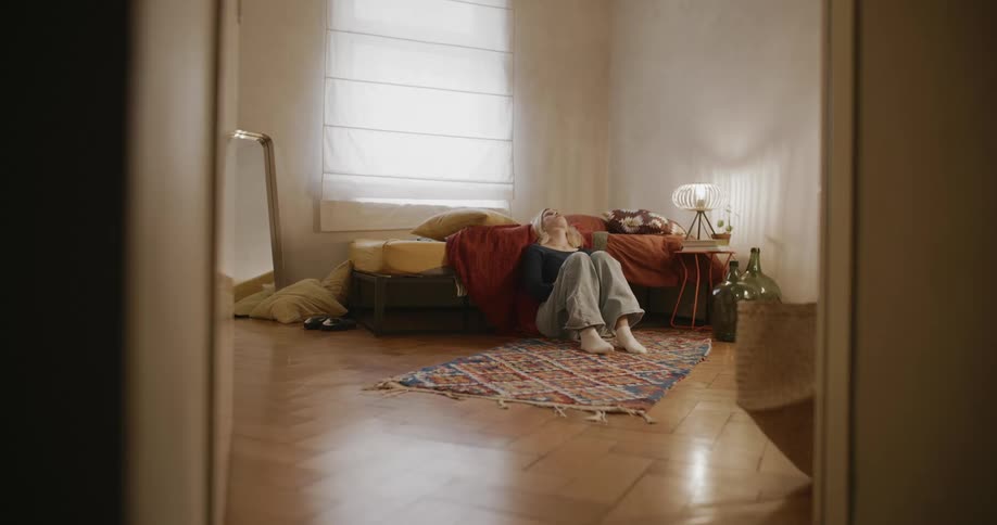 Young woman sitting on floor leans her head back against bed after looking at smartphone
